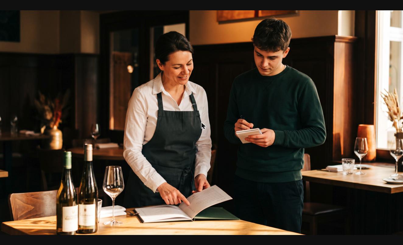 Erfahrene Servicekraft erklärt einem neuen Mitarbeiter im Restaurant freundlich die Weinkarte.